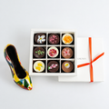 Box of chocolates with floral designs next to a colorful high-heeled shoe on a white background